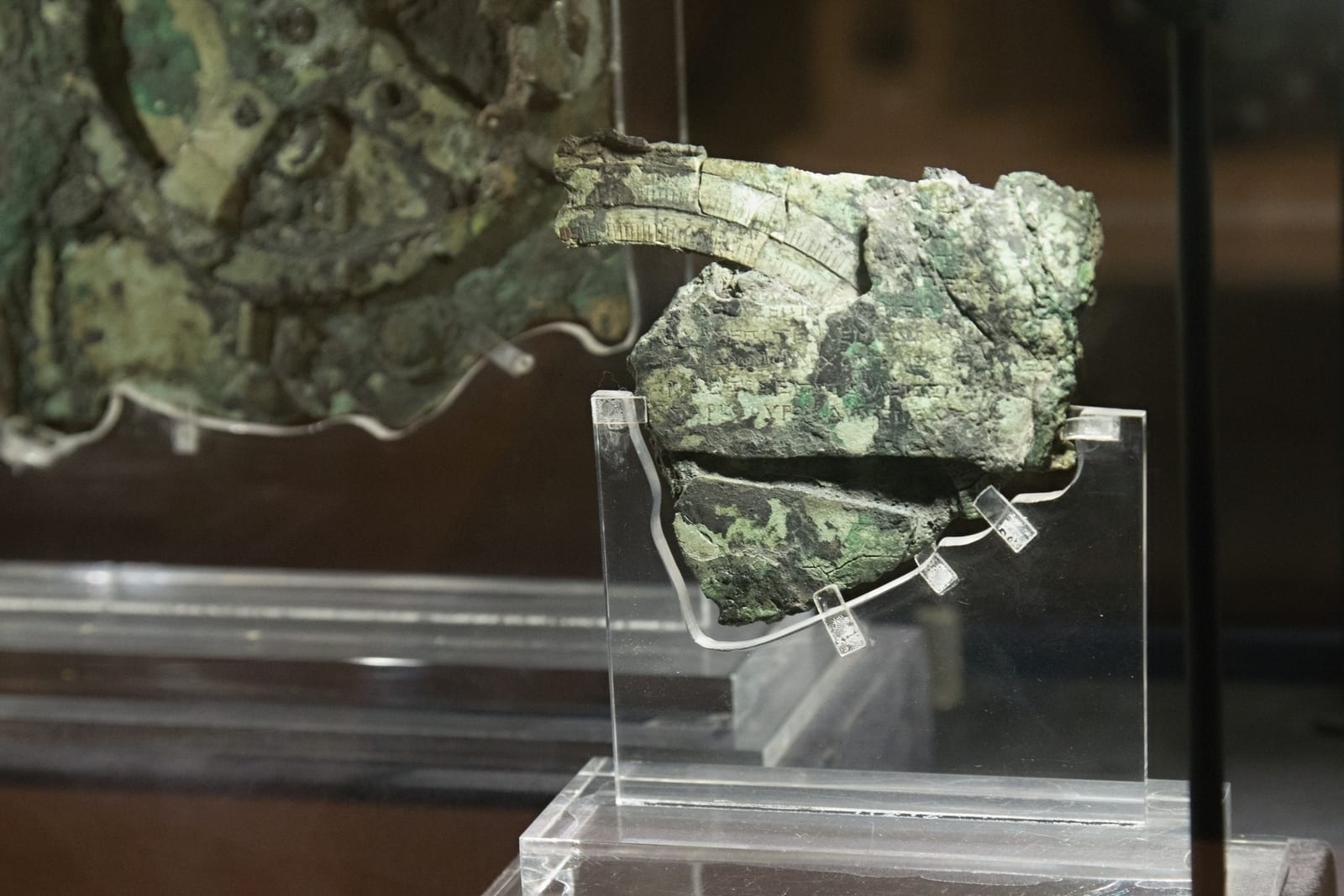 Fragment B of the Antikythera mechanism on display at the National Archaeological Museum in Athens, showing a curved graduated dial with fine tick marks at the top and visible ancient Greek inscriptions engraved on the corroded bronze surface, which served as part of the device's user manual. Photo: Zde, CC BY-SA 4.0.