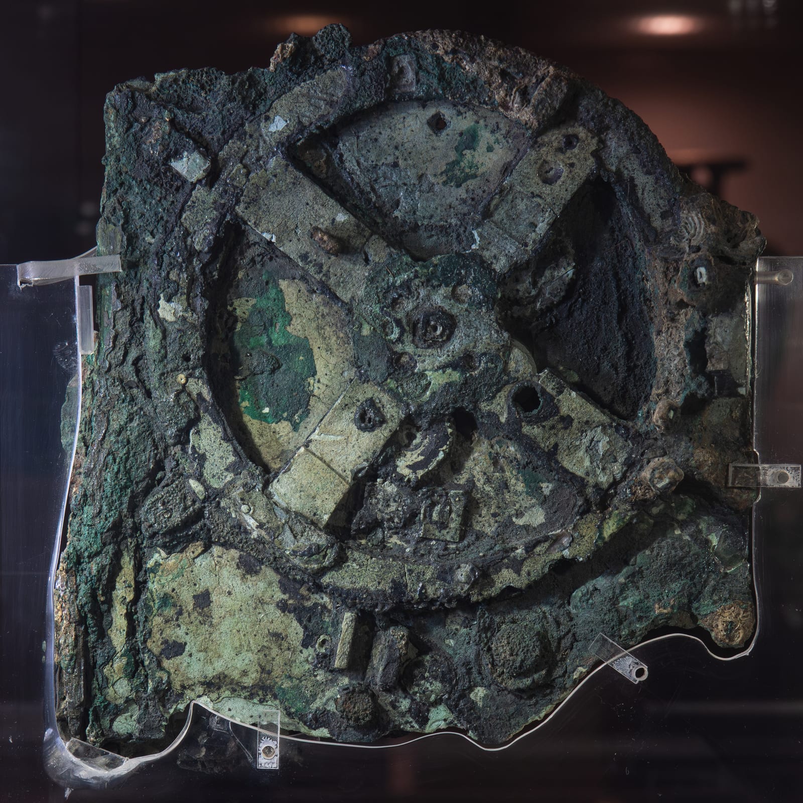 Fragment A of the Antikythera mechanism, the largest surviving piece: a dense corroded mass of green and brown bronze dominated by the four-spoked main drive gear with its triangular teeth, with parts of at least 27 other gears embedded around it, mounted in a clear acrylic frame. Photo: Logg Tandy, CC BY 4.0.