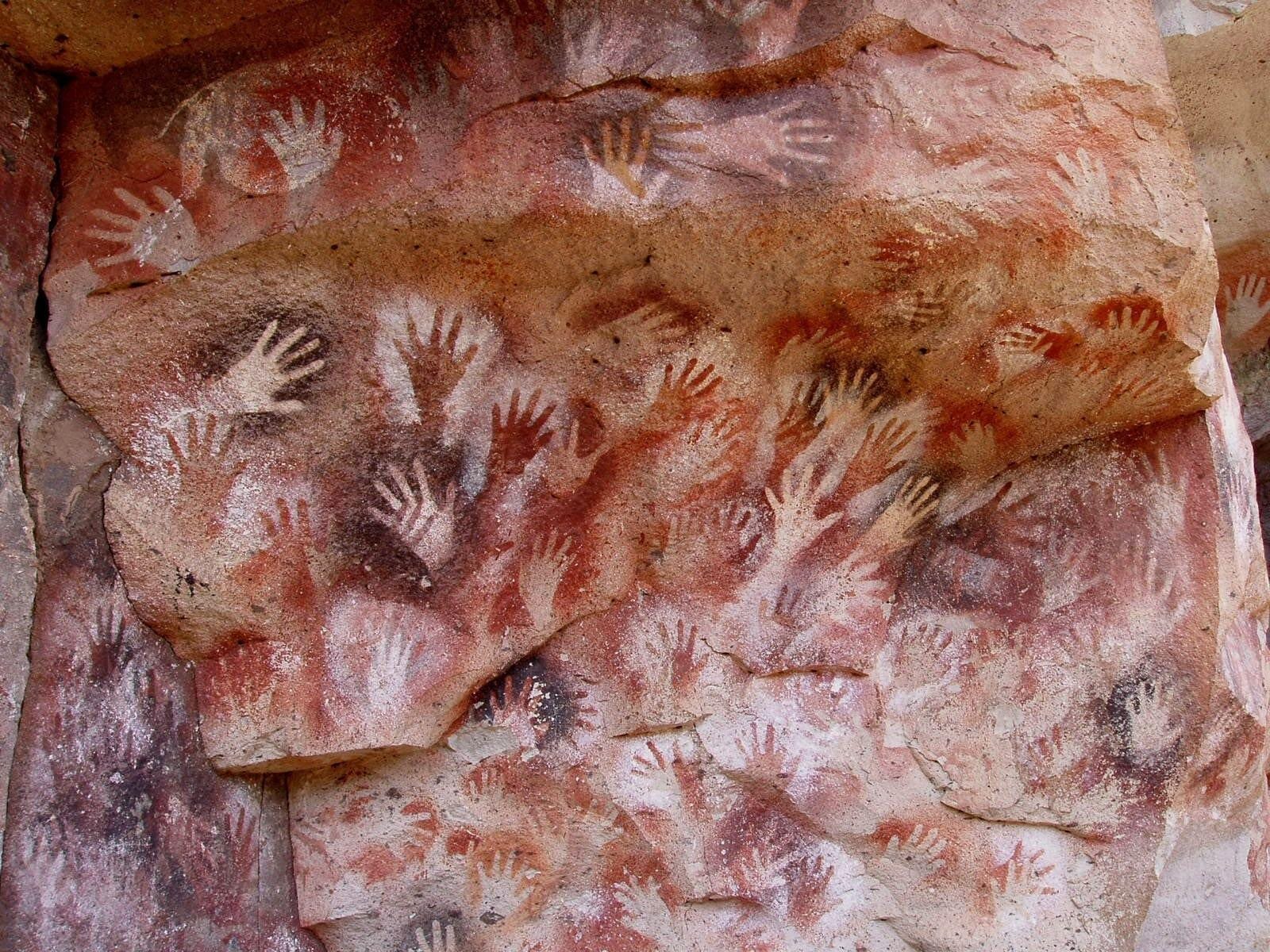 Cave wall covered in hand stencils at Cueva de las Manos in Argentina, showing dozens of overlapping hand outlines in red, black, and white.