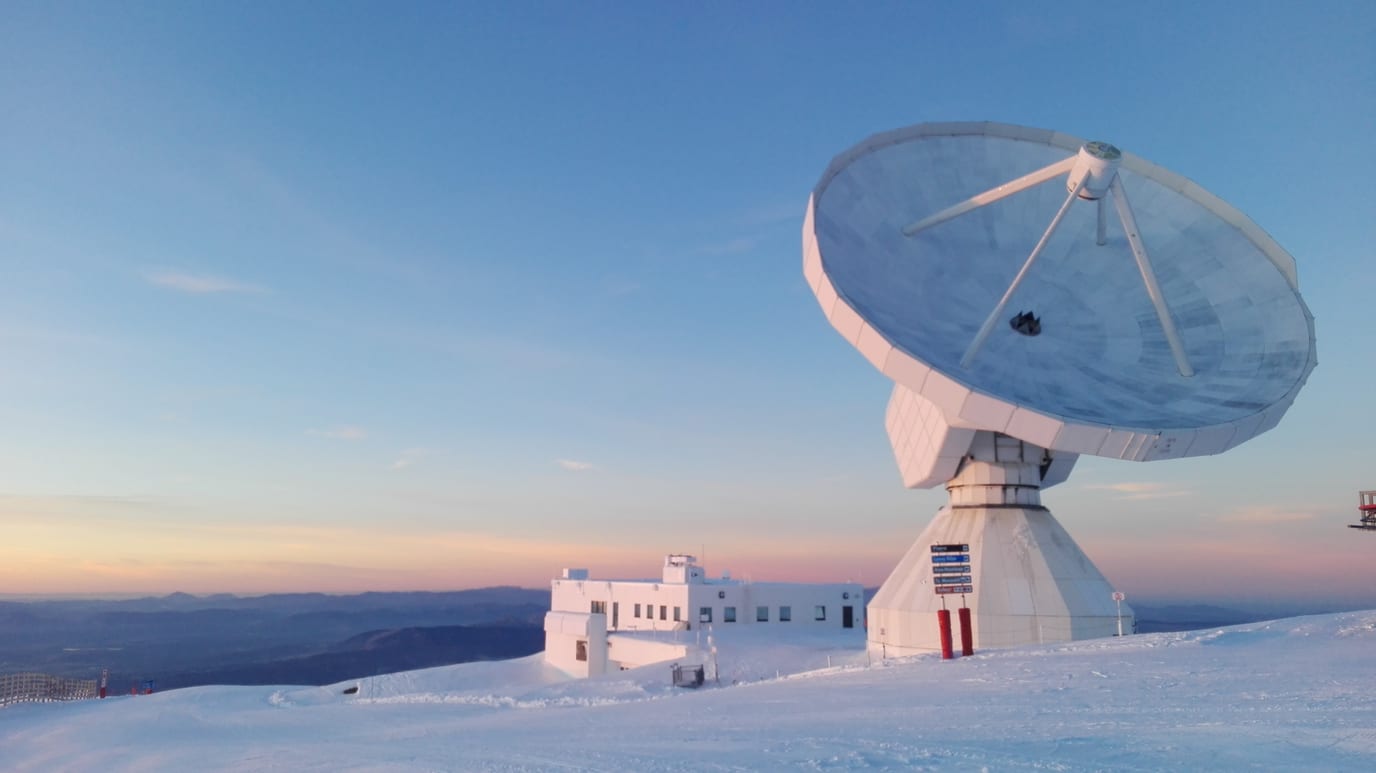 The white dish of the IRAM 30-meter radio telescope on Pico Veleta in Spain's Sierra Nevada, seen against a twilight sky over snowy mountains.