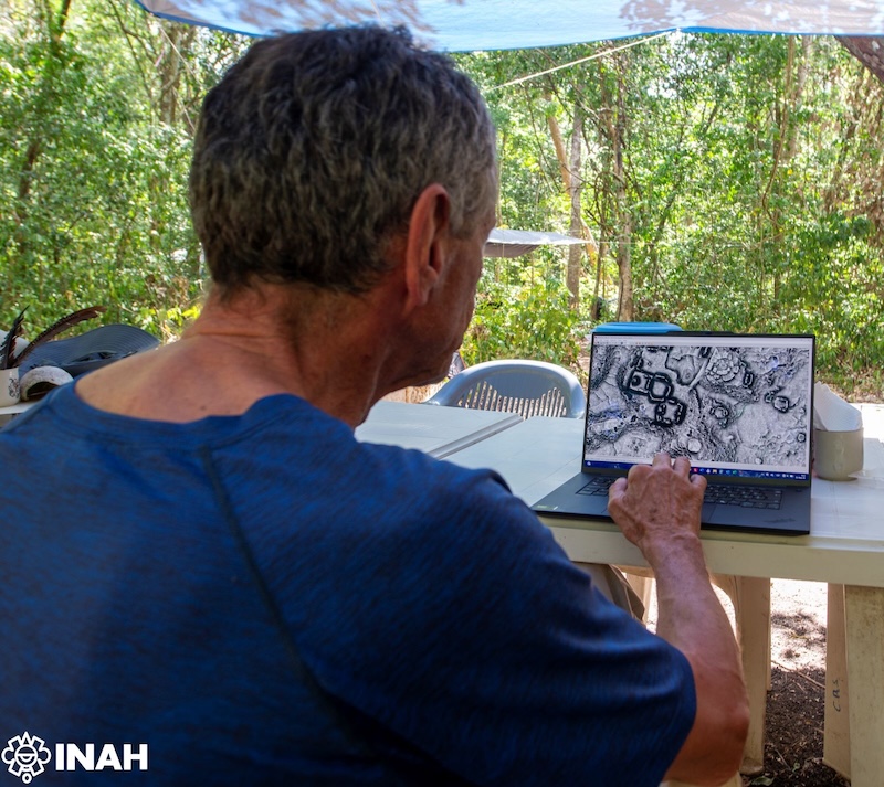 An archaeologist at the Minanbé field camp studies LiDAR data on a laptop under a blue tarp, surrounded by jungle.