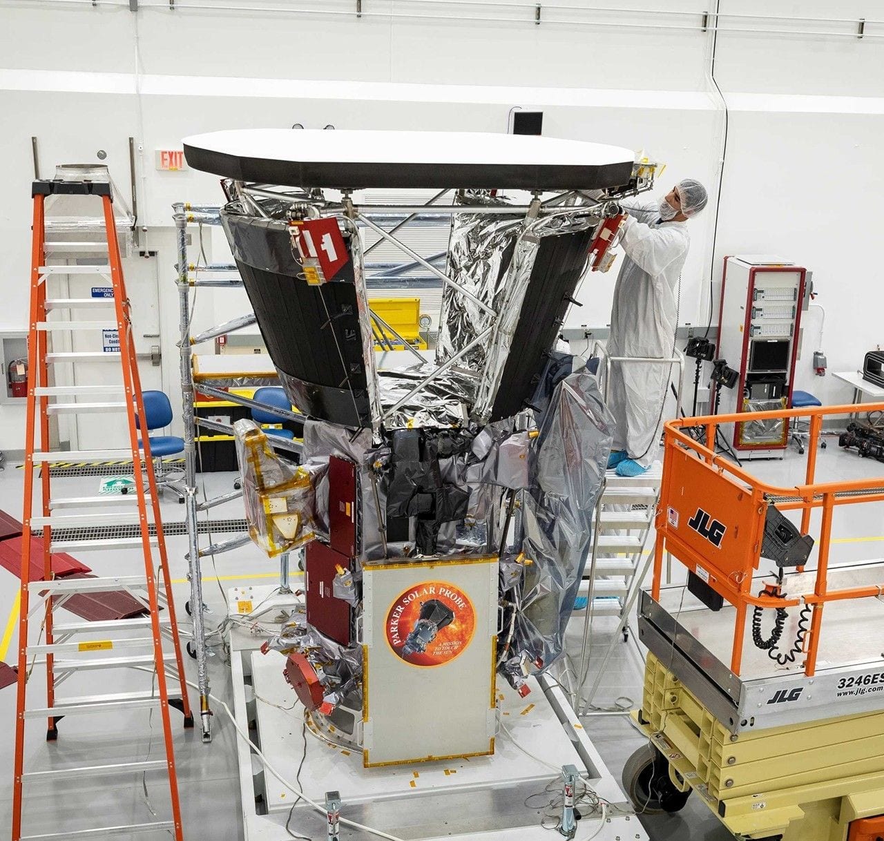 A technician in a white cleanroom suit standing on a ladder, working on the Parker Solar Probe spacecraft just below its large circular white heat shield.