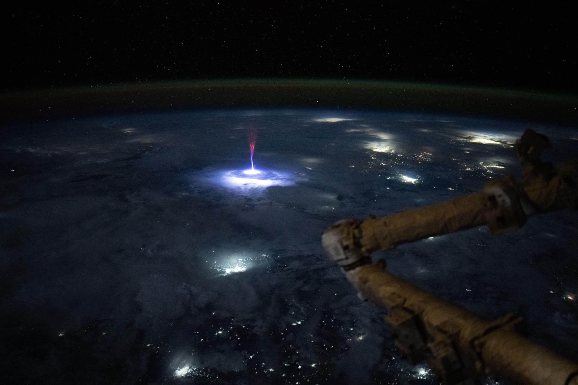 Photograph from the International Space Station at night: a bright blue-white lightning flash inside a storm cloud below, with a tall reddish-purple gigantic jet of light shooting upward from the storm top into a black, star-filled sky, a thin green airglow along the horizon, and part of the station visible in the foreground. Credit: NASA / Nichole Ayers.