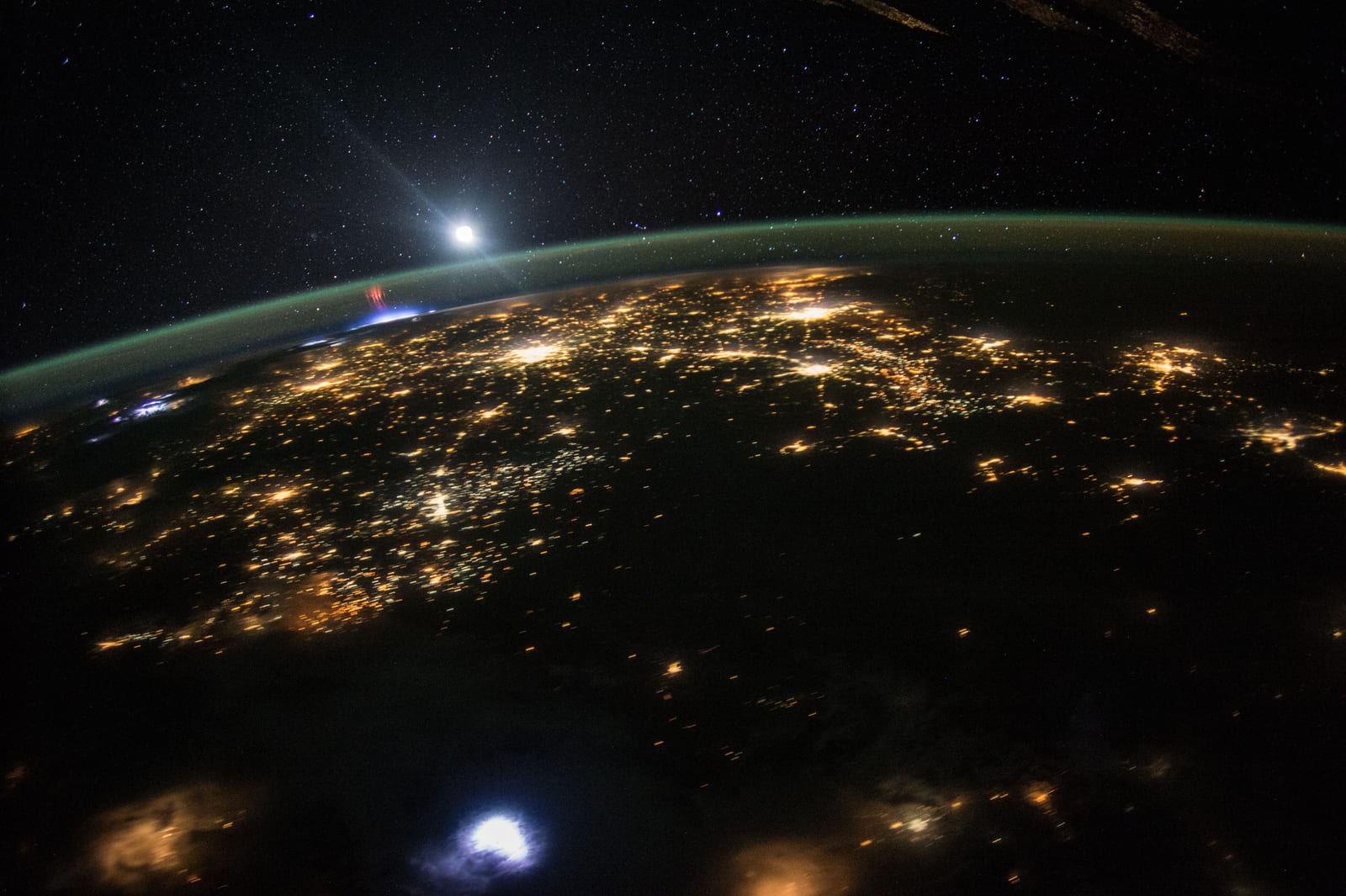 Astronaut photograph of Earth at night: a sweep of golden city lights below, a thin green airglow along the curved horizon, and a small cluster of red sprites visible above a distant storm near the top left edge of the image. Credit: NASA / Expedition 44 crew.