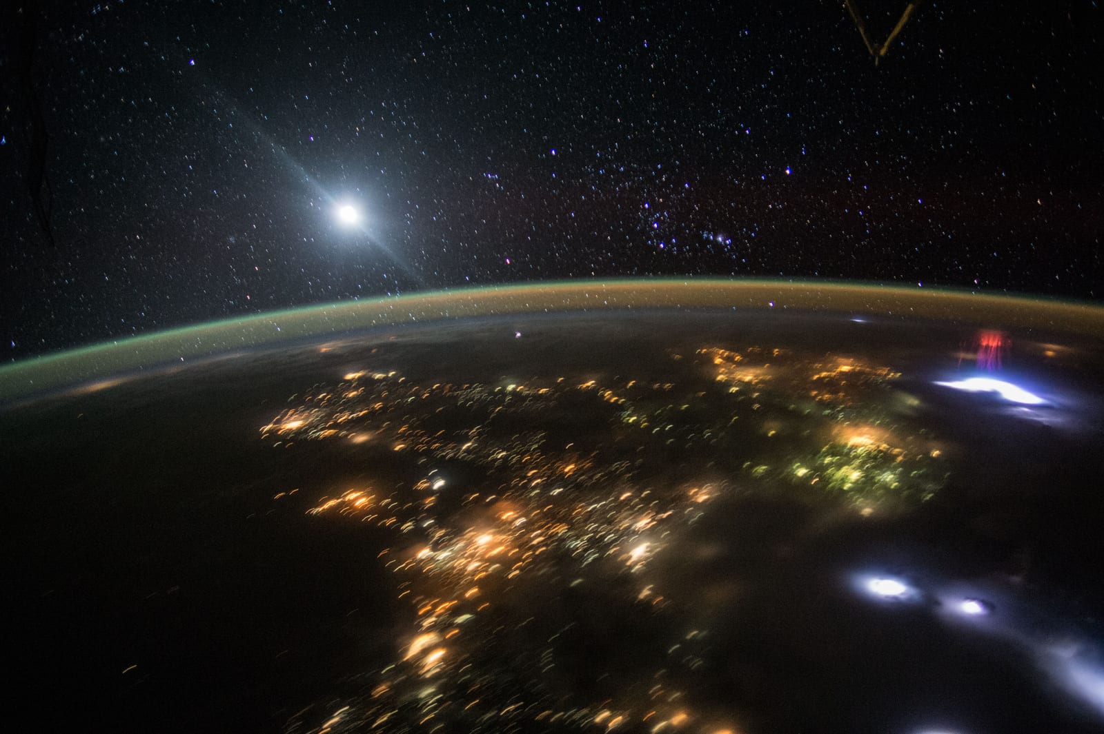 Astronaut photograph of Earth at night from the International Space Station: city lights glow gold below, a bright white-blue lightning flash illuminates a storm cloud on the right, and a cluster of red jellyfish-shaped sprites glows directly above it against the black sky. Credit: NASA / Expedition 44 crew.