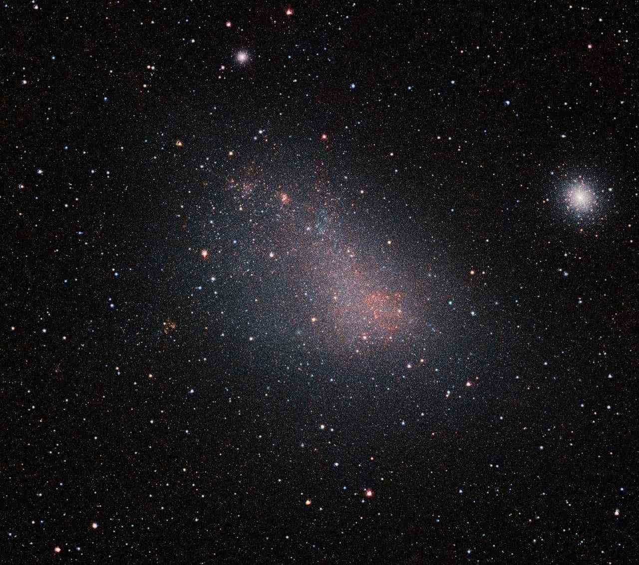 An infrared view of the Small Magellanic Cloud galaxy captured by ESO's VISTA telescope, showing millions of individual stars packed densely across the irregular galaxy against a dark background. The right side of the image contains the bright globular cluster 47 Tucanae.