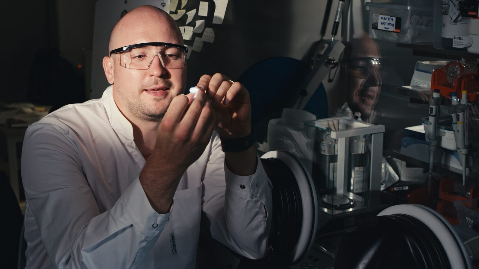 A sodium-ion battery researcher in a white lab coat and safety glasses holds a small cylindrical button-cell battery between his fingertips in a dim laboratory, a bright white LED glowing from the cell and illuminating his face and hands, with a glovebox workstation and lab supplies behind him. Photo: Tavo Romann, CC BY 4.0, via Wikimedia Commons.