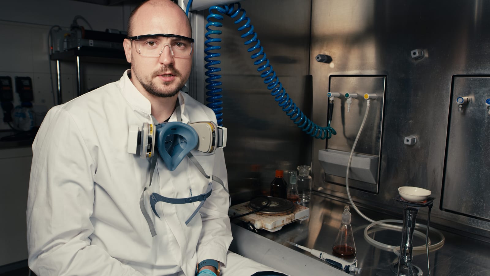 A sodium-ion battery researcher in a white lab coat and safety glasses sits in front of a stainless steel fume hood, preparing cathode material on a hot plate with a glass flask of dark liquid, a pipette, and an evaporating dish, with gas and vacuum lines on the back wall. Photo: Tavo Romann, CC BY 4.0, via Wikimedia Commons.