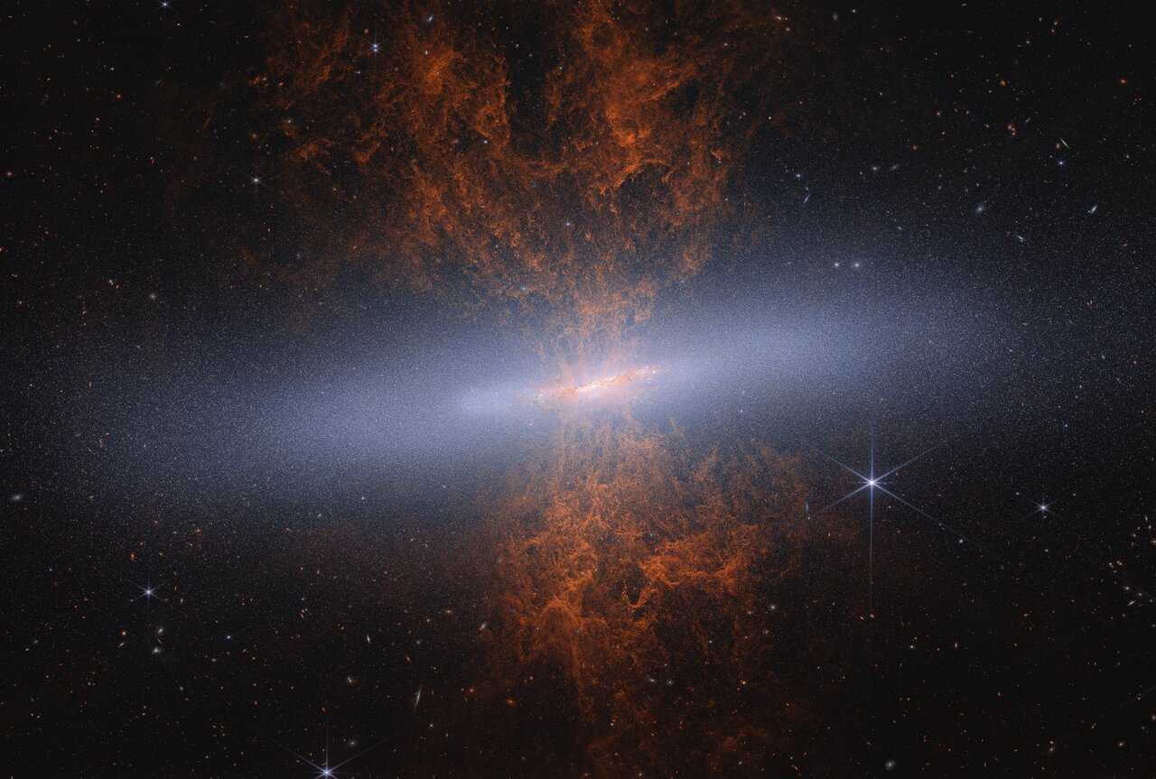 Webb NIRCam image of the Cigar Galaxy showing a bright central disk packed with millions of tiny blue-white stars against black space, with the signature hourglass-shaped outflow plumes in yellow and orange